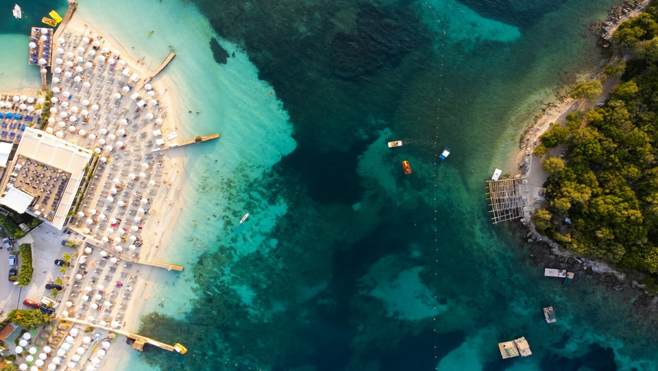 Vista aerea di una spiaggia affollata con acqua turchese e una piccola isola.