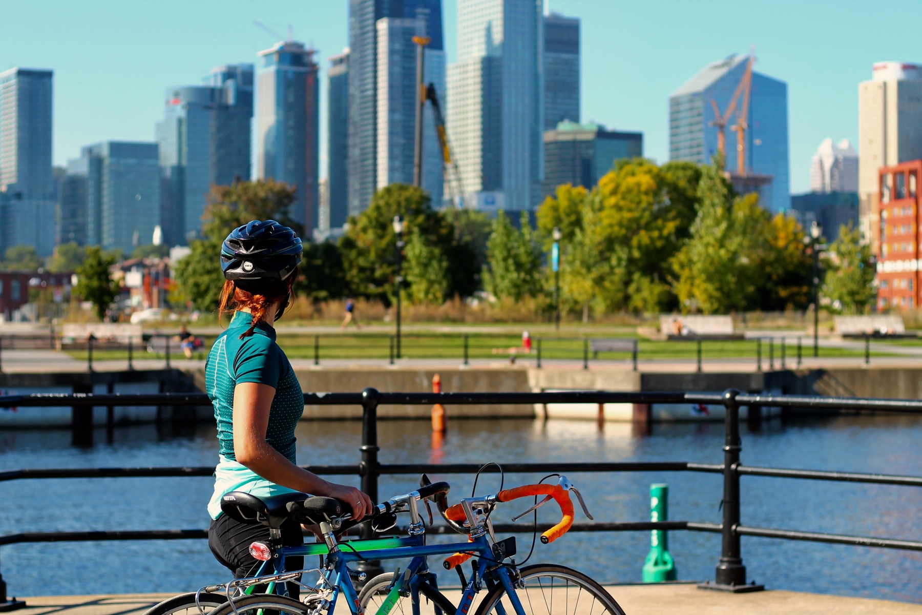 woman biking along the Lachine Canal with a view of downtown Montreal on a sunny day