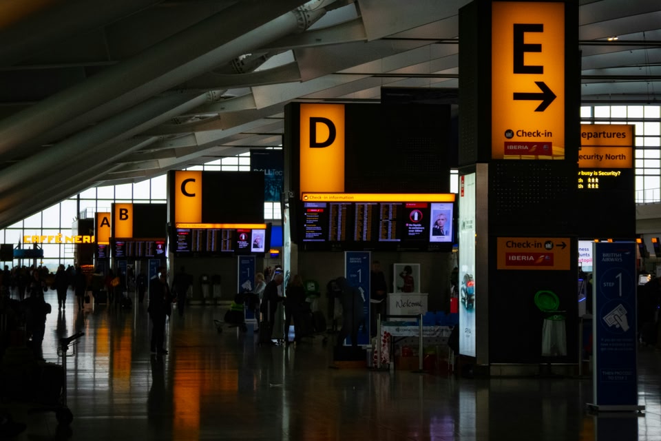 Large, glowing yellow signs labeled A through E hang above check-in counters and flight information screens in a modern, spacious airport terminal.