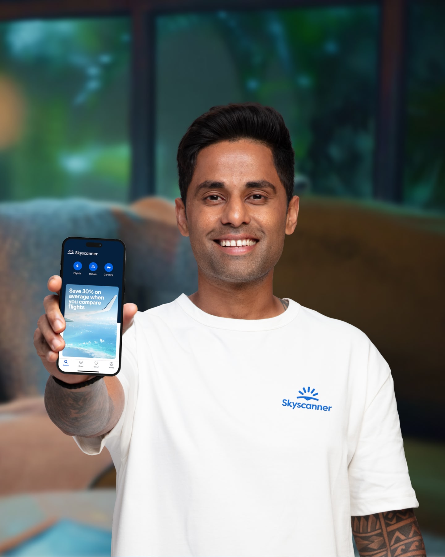 Indian cricketer, Suryakumar Yadav, holds a smartphone showing the Skyscanner app up to the camera, smiling.