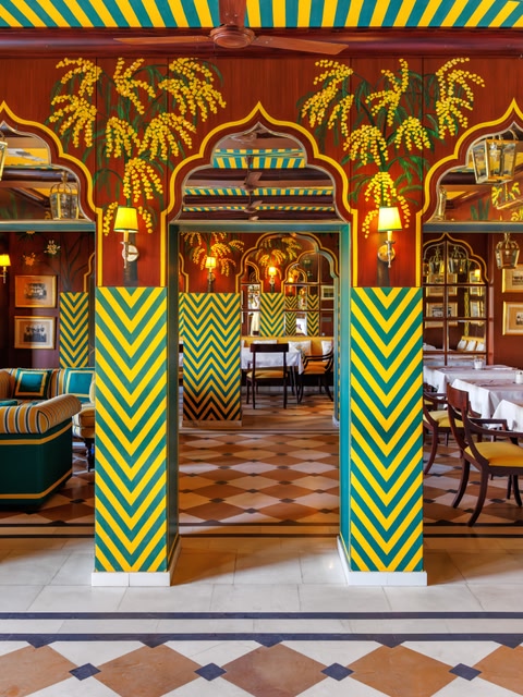 The vibrant interior of Polo Palladio restaurant in Jaipur, featuring boldly painted Mughal-style arched columns in green, yellow, and blue chevron stripes, with painted palm tree murals and richly decorated ornate decor.