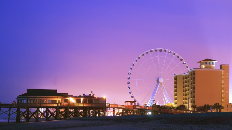 A scenic view of Myrtle Beach at sunset, featuring a large Ferris wheel, illuminated buildings, and a pier extending into the ocean.