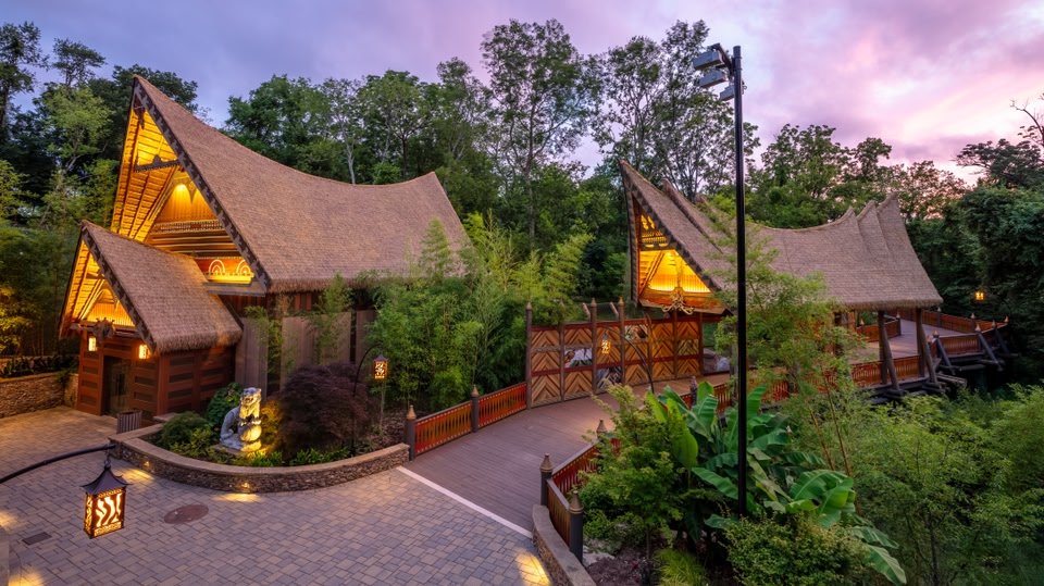 Illuminated resort-style buildings with thatched roofs set in a lush, tree-filled environment at dusk.