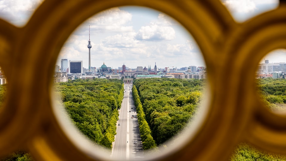 A circular, framed view from the top of the Victory Column in Berlin, showing a long tree-lined boulevard leading to the city skyline with the Fernsehturm television tower prominently visible.