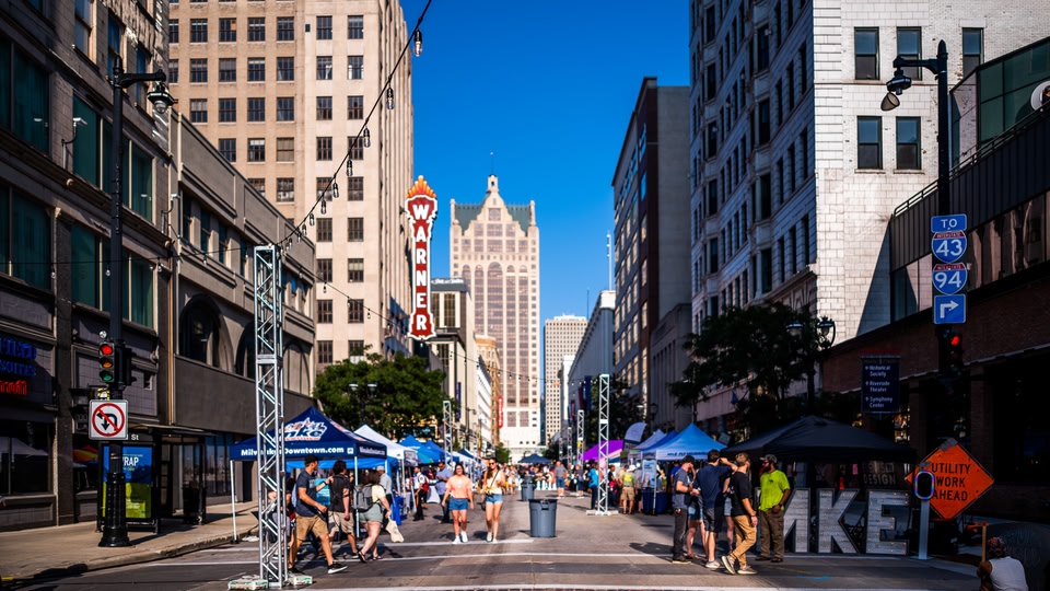 A busy city street with people walking and standing by market stalls during an event.