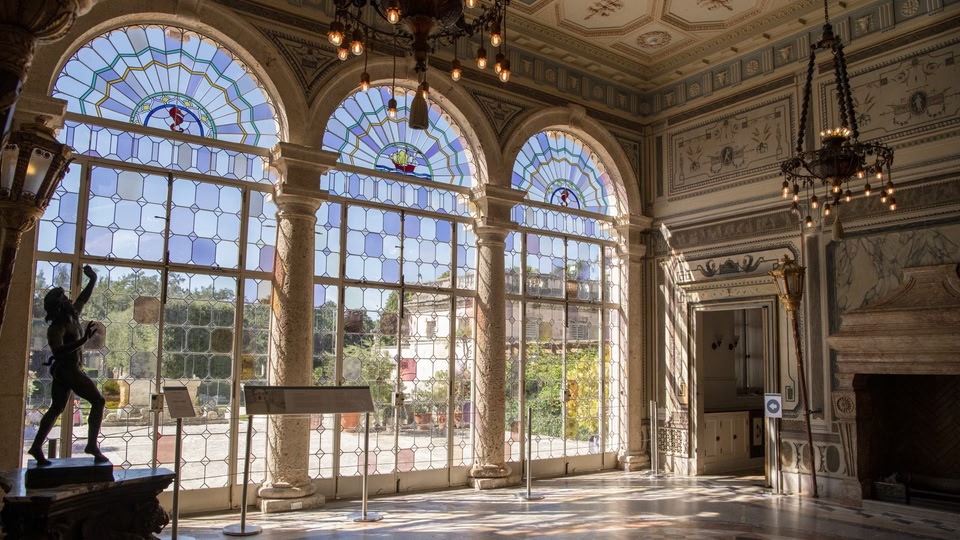 An ornate interior room with large stained-glass windows, detailed ceiling, and classical architecture. A statue and informational stand are present.