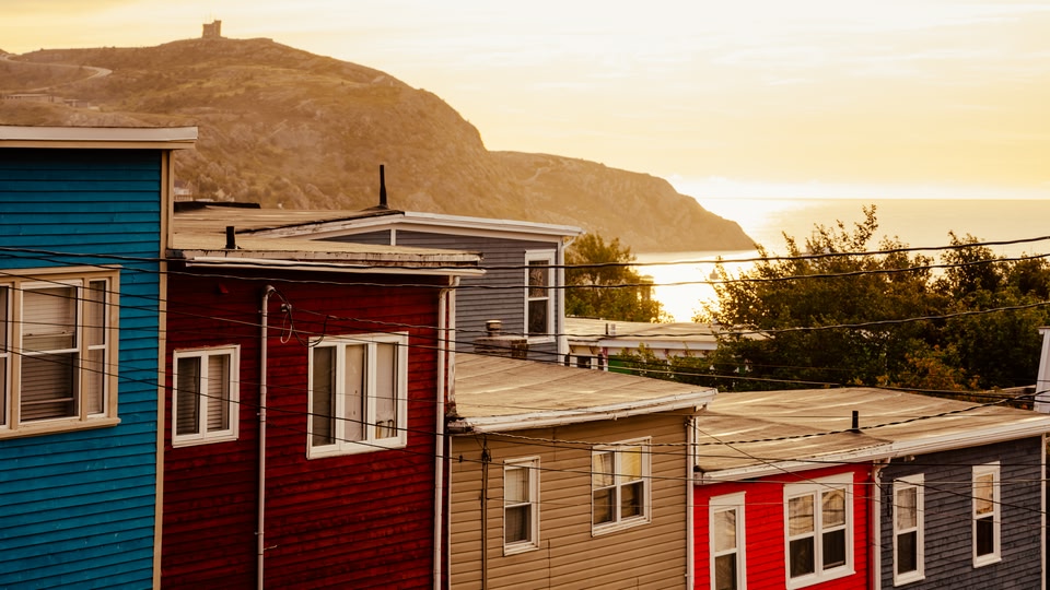A row of colorful houses with the sun setting behind a mountain and the sea in the distance.