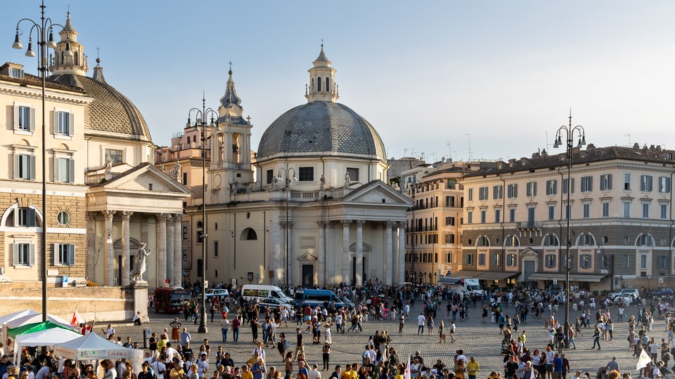 A large crowd of people in Piazza della Repubblica, Rome, with historic buildings and domes in the background.