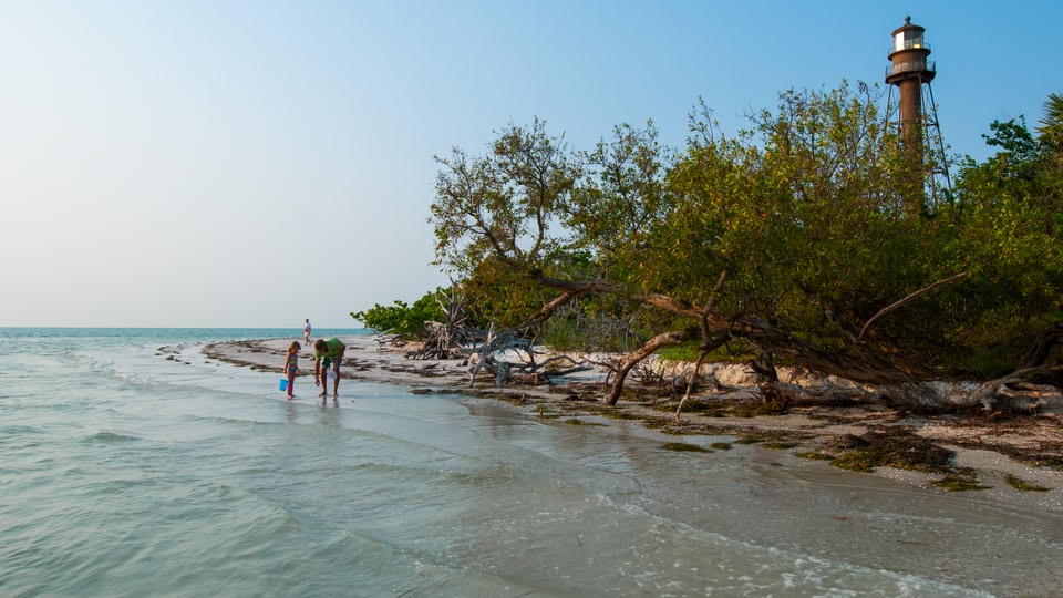 Three people, including a woman with a child, exploring a sandy beach near a lighthouse in Fort Myers, Florida.