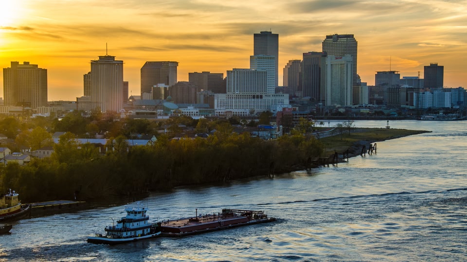 A panoramic view of the New Orleans skyline at sunset with a river in the foreground.
