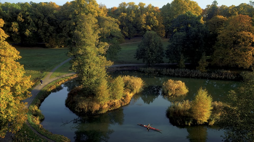 Aerial view of a serene lake surrounded by trees with vibrant autumn colors, with a person in a kayak and another walking on a path.