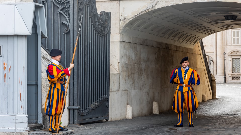 Two Swiss Guards in traditional colorful uniforms standing at attention in front of a gate at St. Peter's Basilica in Rome.