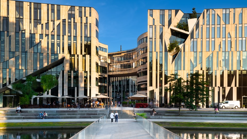Modern architectural buildings with people sitting and walking around, adjacent to a calm body of water.