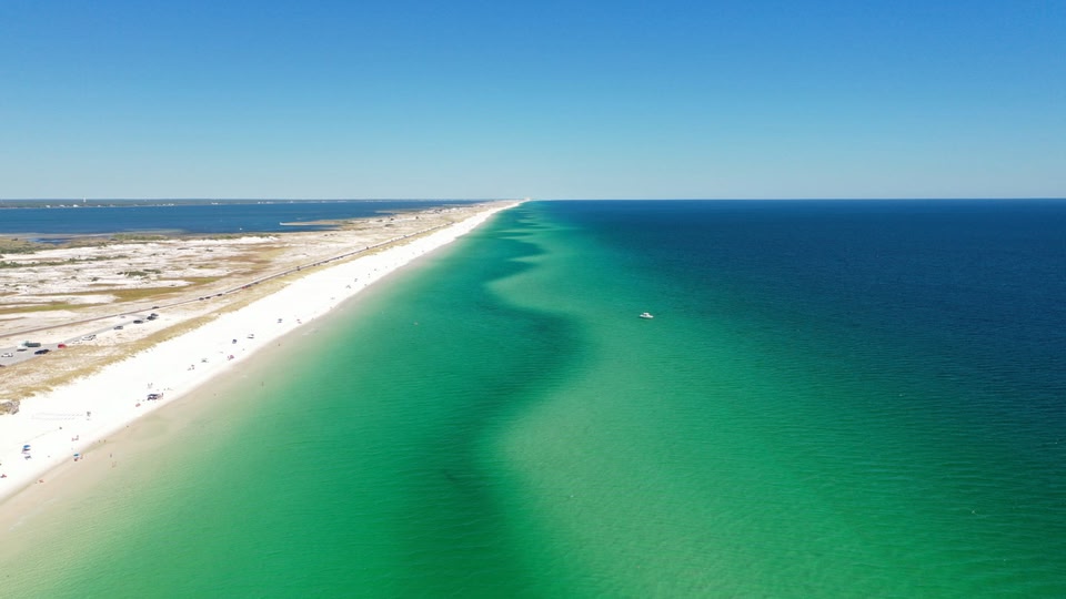 Aerial view of a long sandy beach with clear turquoise water and a busy road along the shore.
