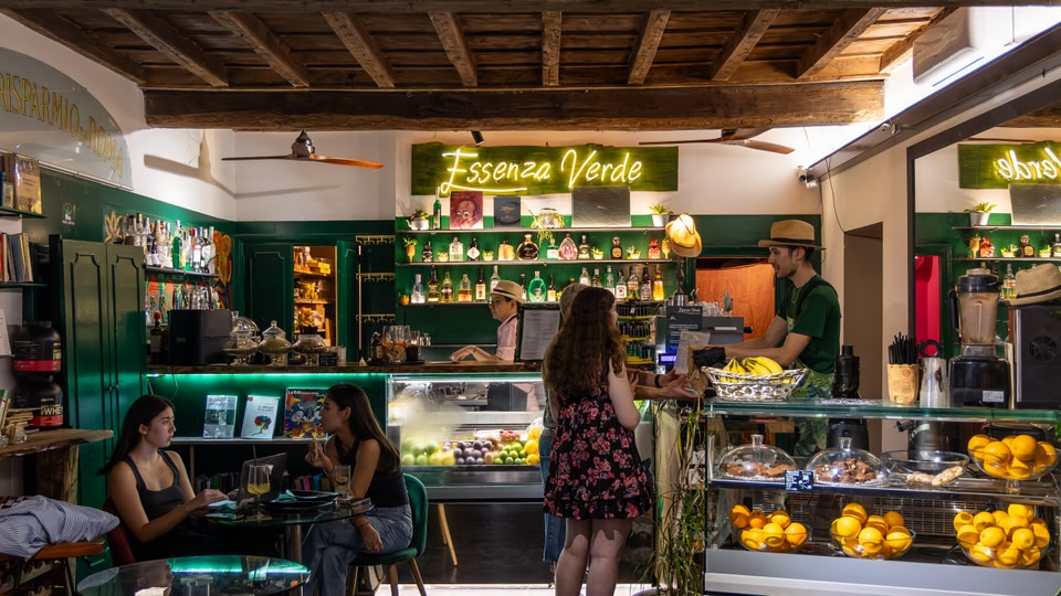 A lively indoor café with patrons, visible food displays, and a bartender serving at the counter.