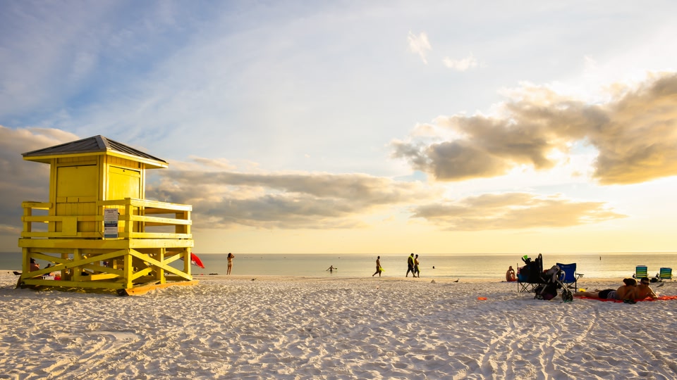 A sandy beach with people walking and relaxing on a cloudy day near a yellow lifeguard station.