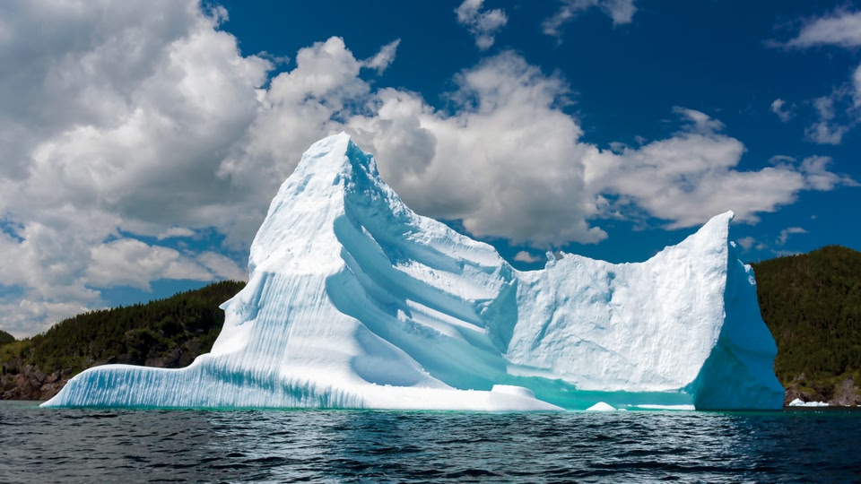 A massive iceberg floating in the ocean with a rocky coastline in the background.