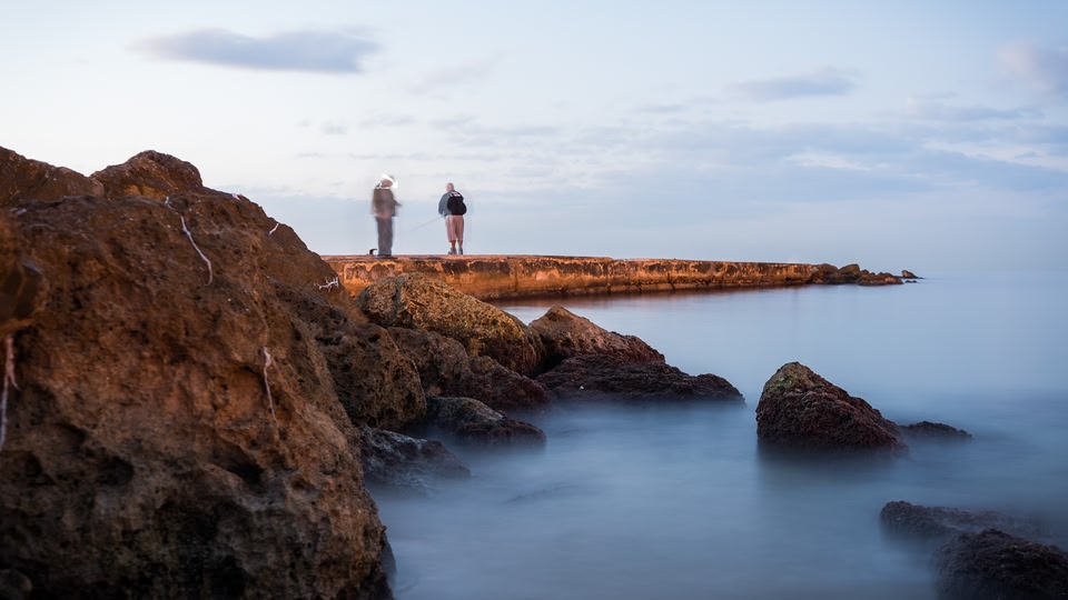 Two people standing at the edge of a rocky pier, with the ocean stretching out behind them during sunset.