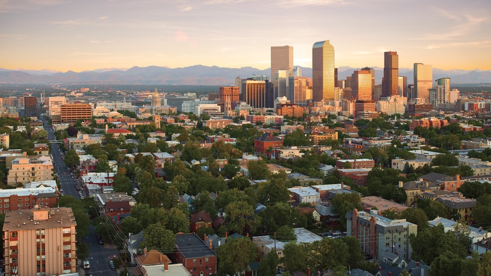 Aerial view of the Denver, Colorado skyline at sunset with a mix of modern and historic buildings, surrounded by greenery and distant mountains.