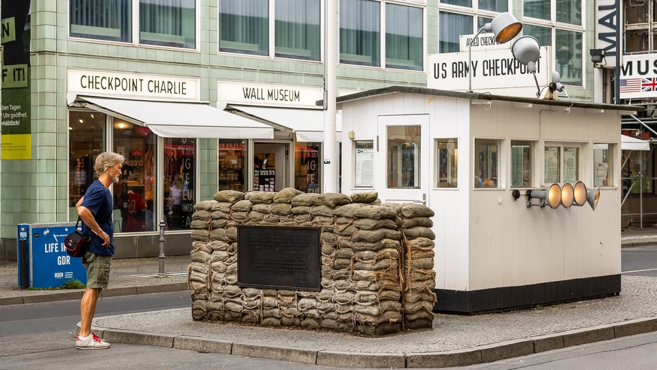 A man walks past the Checkpoint Charlie historical site in Berlin, with a view of the guardhouse and the iconic sandbag barrier.