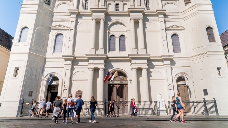 People walking past a large, historic white building with multiple arched windows and doors in New Orleans, Louisiana.