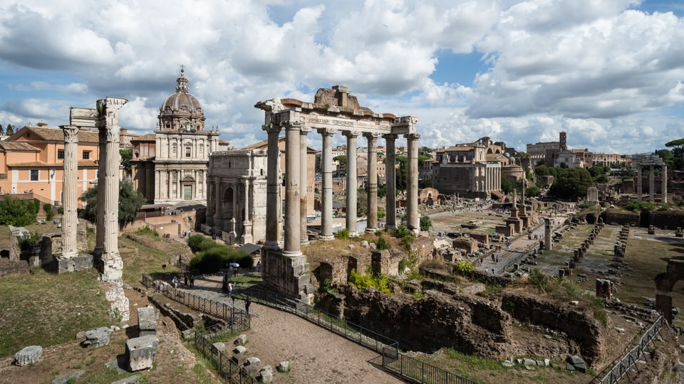 Aerial view of the Roman Forum ruins in Rome, showcasing ancient architecture and columns under a cloudy sky.