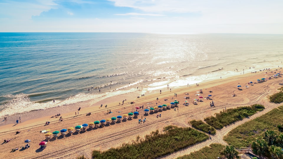 A wooden boardwalk leading through grassy dunes towards a beach and pier at sunset.