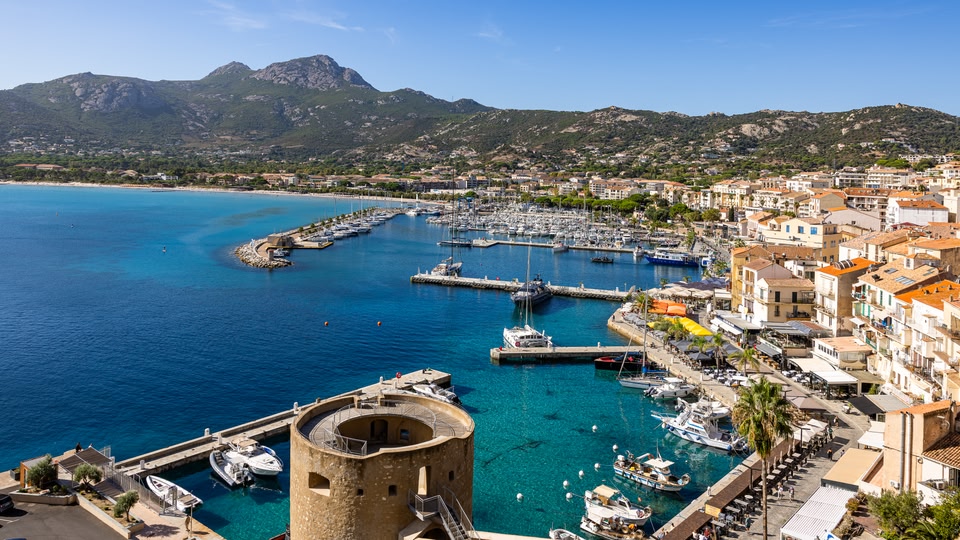 Round stone tower, marina with boats and terracotta-roofed buildings by turquoise sea in Calvi.