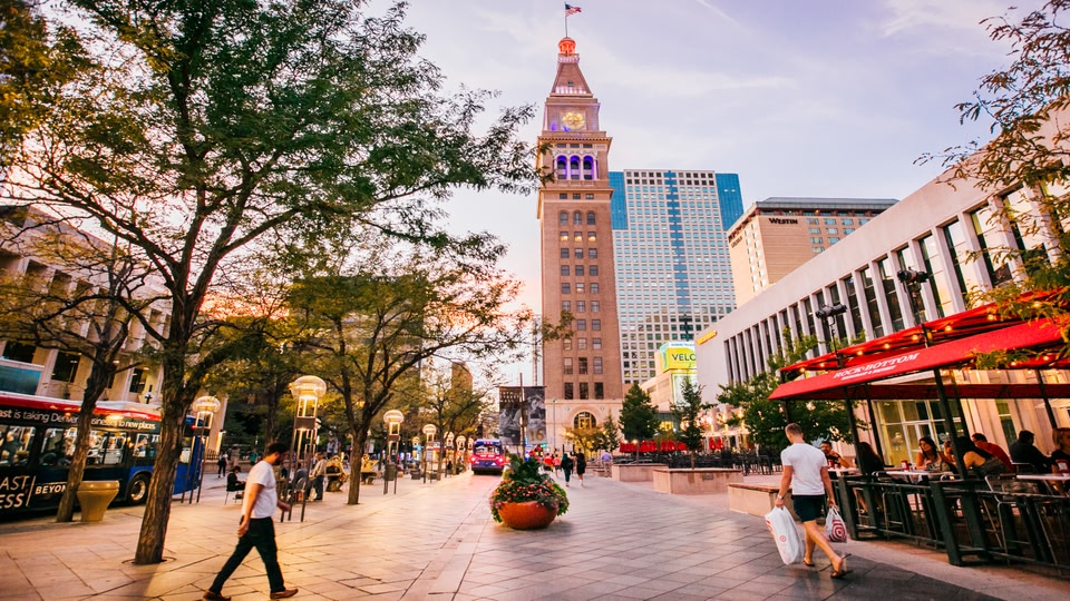 A city street with a prominent clock tower, modern buildings, and people walking around.