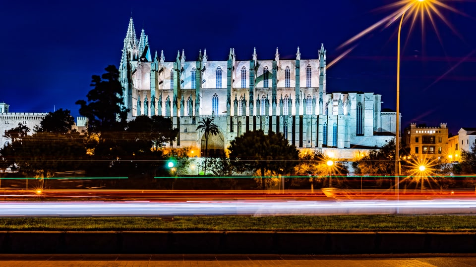 A night view of a large, illuminated cathedral with surrounding buildings and street lights in Palma, Balearic Islands, Spain.