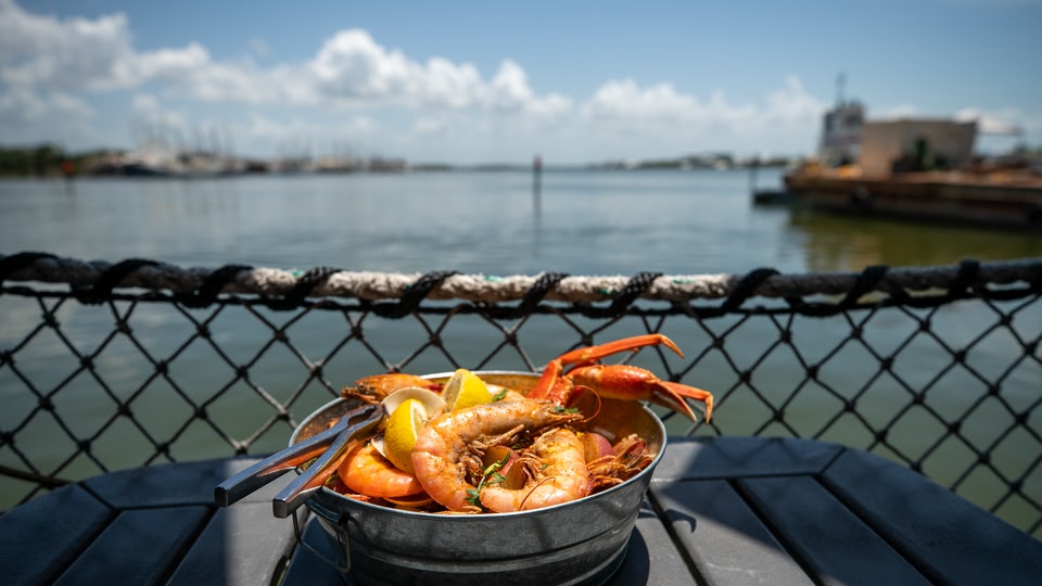 A bowl of shrimp and crab with lemon wedges placed on a table by a waterside fence.