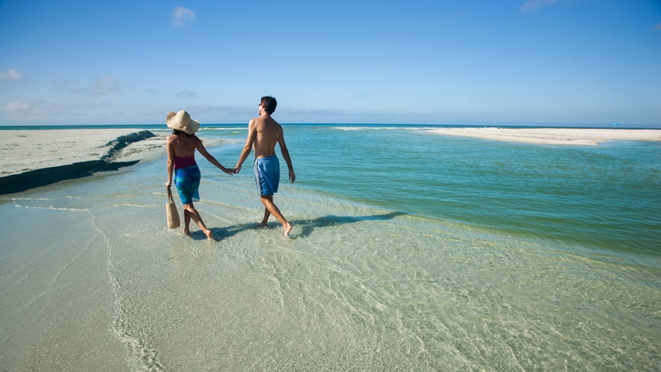 A couple walking hand in hand along a shallow, clear water shoreline at the beach.