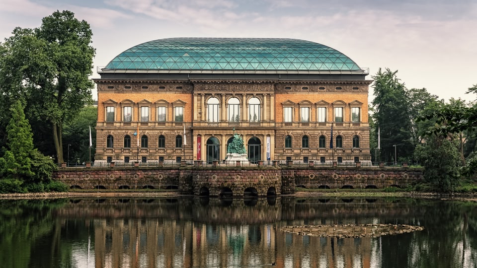 A historic museum building with a glass dome roof, situated near a reflecting pond with lush greenery around it.
