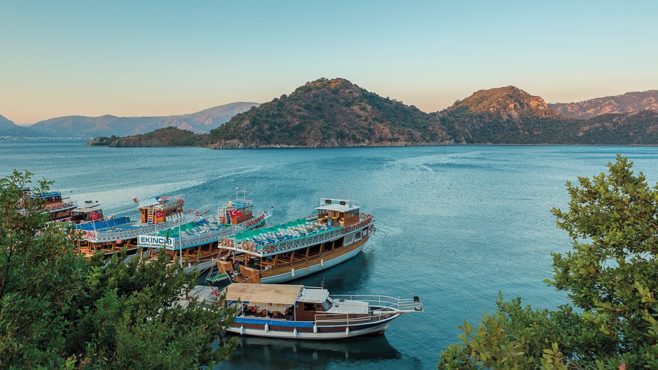 Boats docked along a serene waterfront at sunset, with a large lake and hills in the background.
