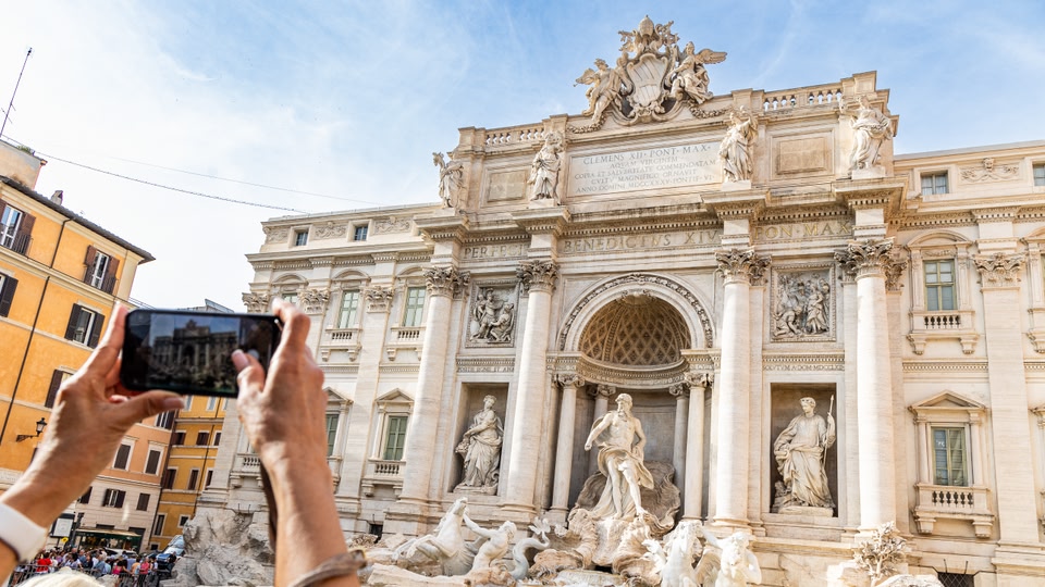 A person is taking a photo of the Trevi Fountain in Rome, a famous baroque fountain with intricate sculptures and a grand design.
