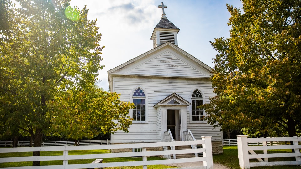An old white church with a cross on top, surrounded by trees and a white picket fence.