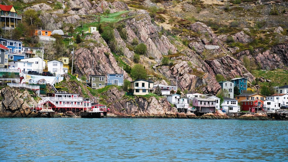 A person stands on a grassy hill overlooking a serene coastal town at sunrise, with the sun shining brightly in the background.