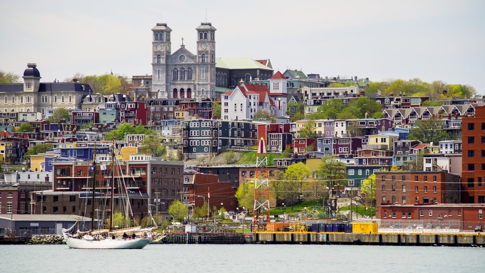 A vibrant waterfront city skyline with colorful buildings and a prominent cathedral, viewed from across the water with a sailboat in the foreground.