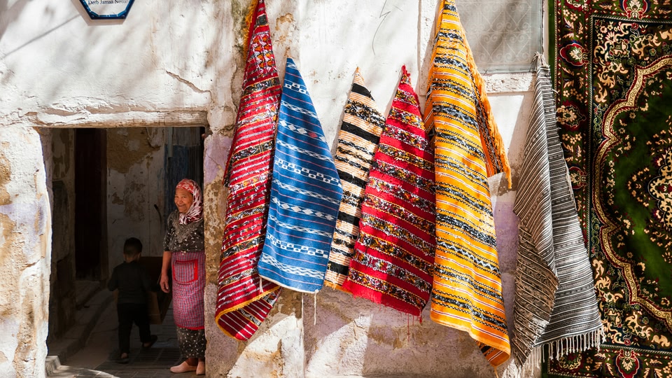 An alleyway stall in Fes with bright hanging rugs and a person standing in a doorway.