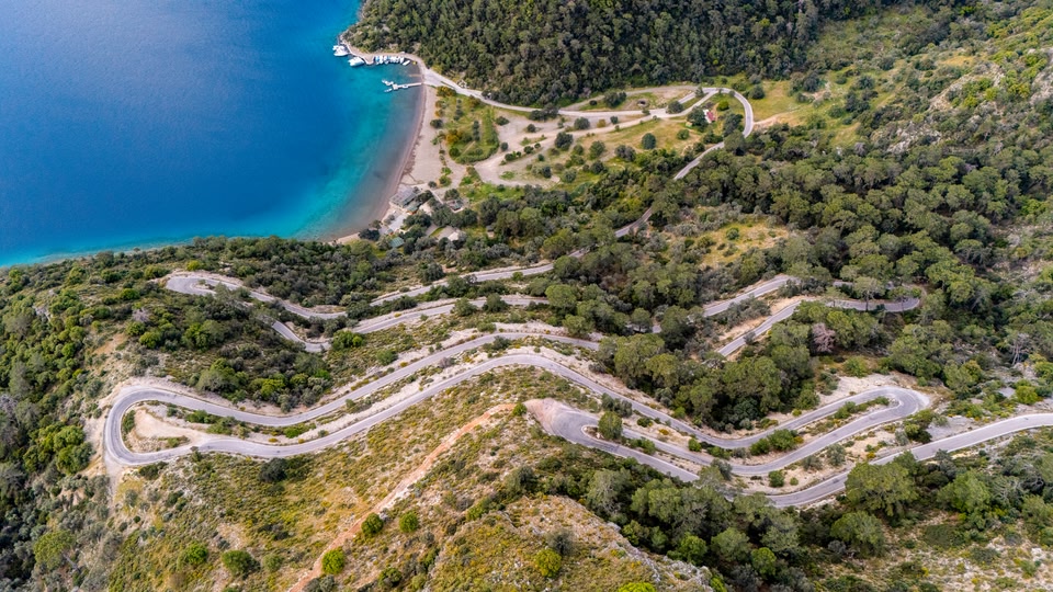 Aerial view of a winding road with a coastal beach and dense forest in the background.