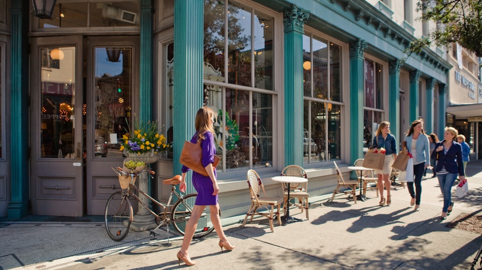 A woman in a purple dress walks down a street lined with shops and cafes on a sunny day.