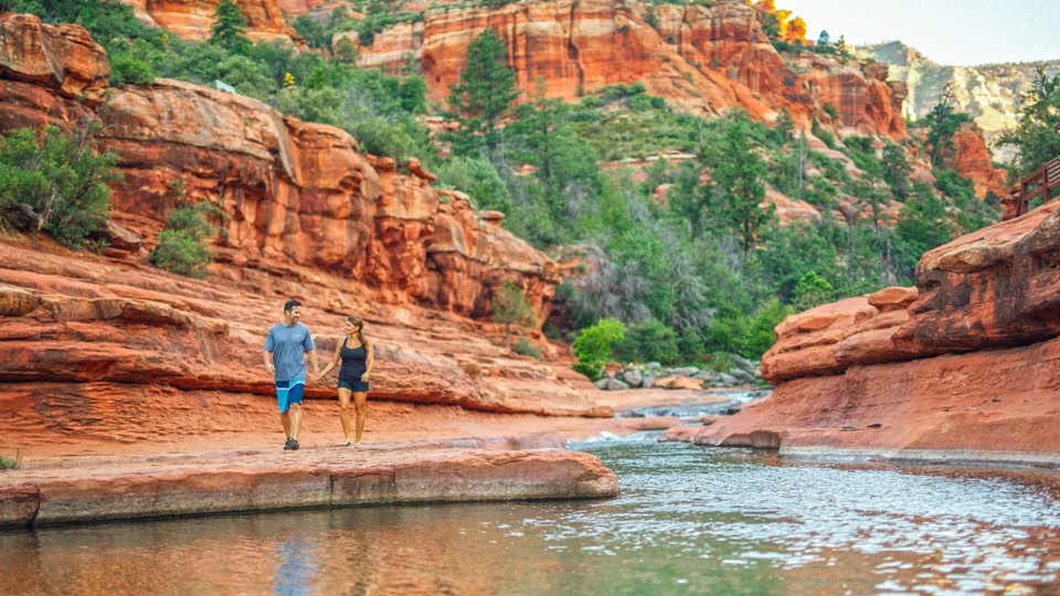 A couple walking along a rocky riverbank in Sedona, Arizona, surrounded by red rock formations and green foliage.