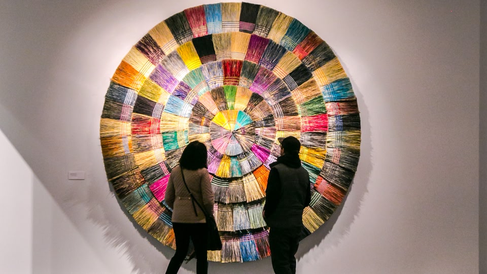 Two people standing in front of a colorful, circular art installation in an art gallery.