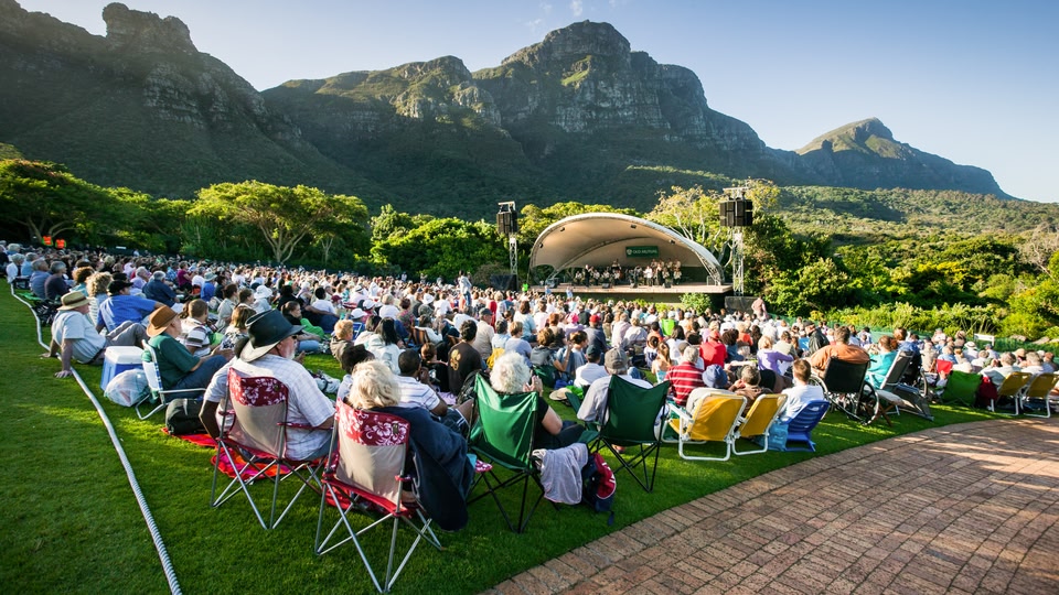 A large crowd of people seated on chairs outdoors, watching a live performance on a stage set against a backdrop of mountains.