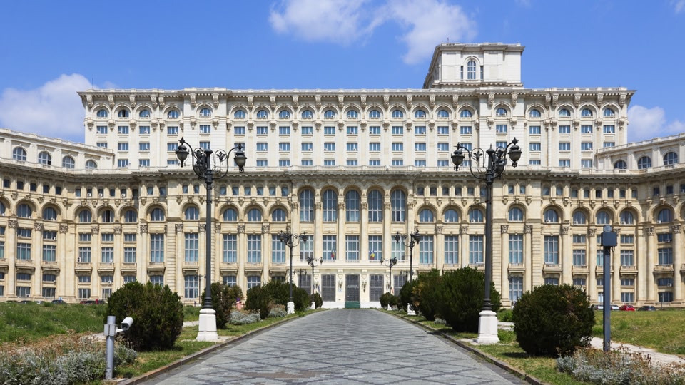 A grand, neoclassical building with many windows and decorative elements, surrounded by greenery and a clear path leading to its entrance.