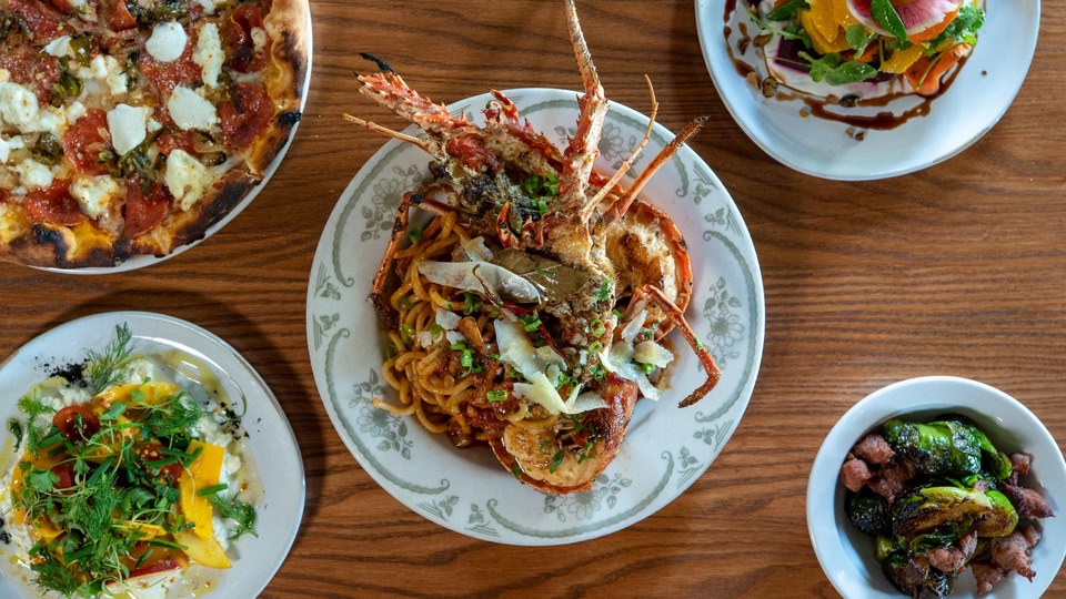 Several plates of food including a lobster dish, salad, pizza, and sides on a wooden table