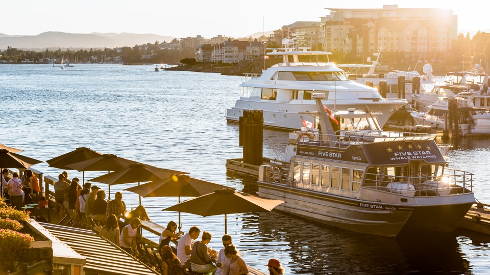 A group of people are socializing and dining outdoors by the waterfront with boats docked nearby at sunset.