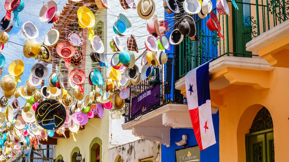 Bright straw and fabric hats suspended over a narrow street with balconies and a Panama flag in Panama City.