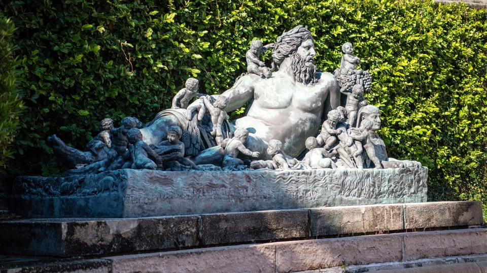 A large stone statue depicting a reclining figure surrounded by children, situated outdoors against a backdrop of trimmed hedges.