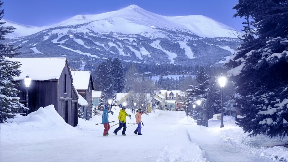 Three skiers walking with their ski gear on a snowy street in Breckenridge, Colorado, with a large snowy mountain in the background.